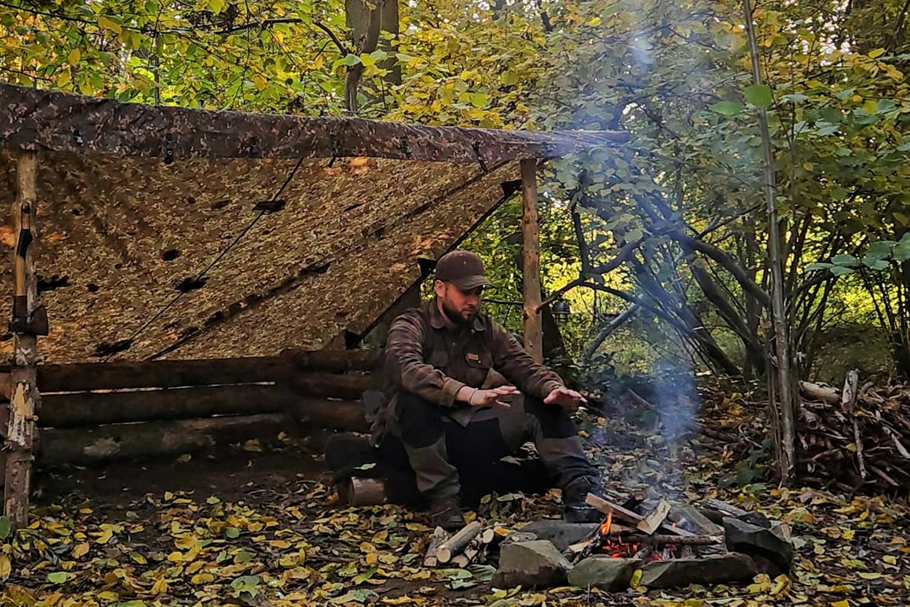 Bushcrafter Cord sitzt im Herbstwald am Feuer vor einem Lean-to-Aufbau des DD Tarp 3x3 in Multicam