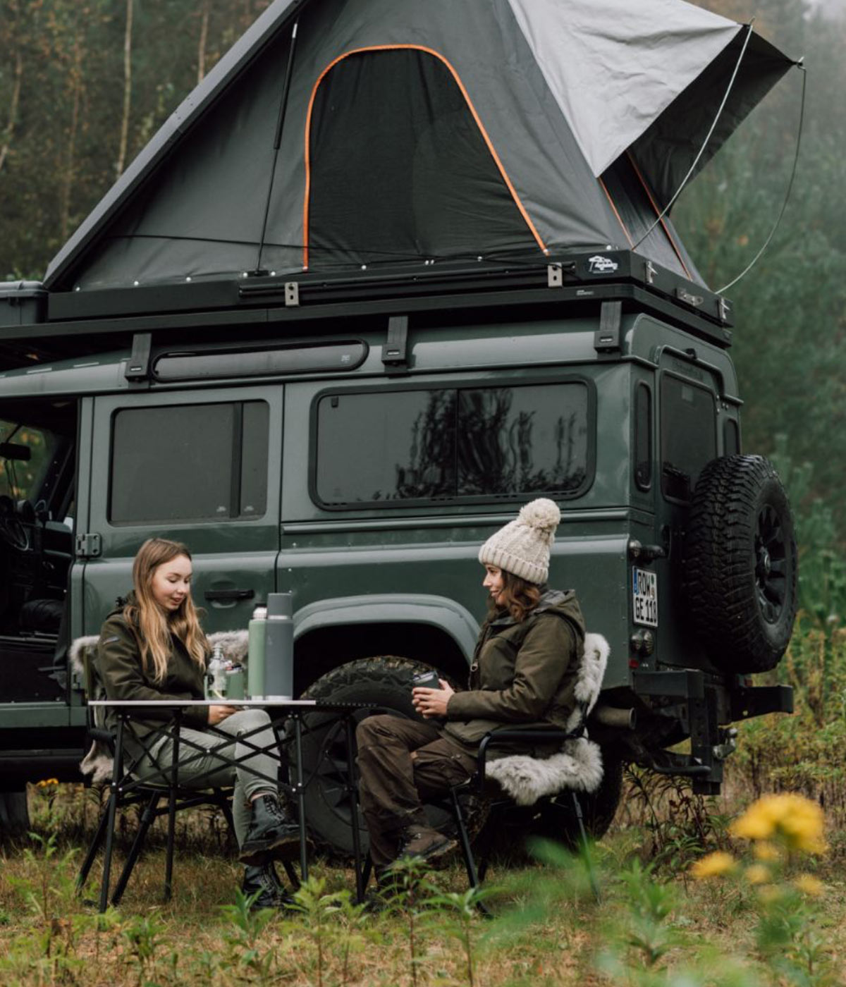 Zwei Frauen sitzen auf Campingstühlen mit Schaffellen vor einem Geländewagen mit Dachzelt im Herbstwald