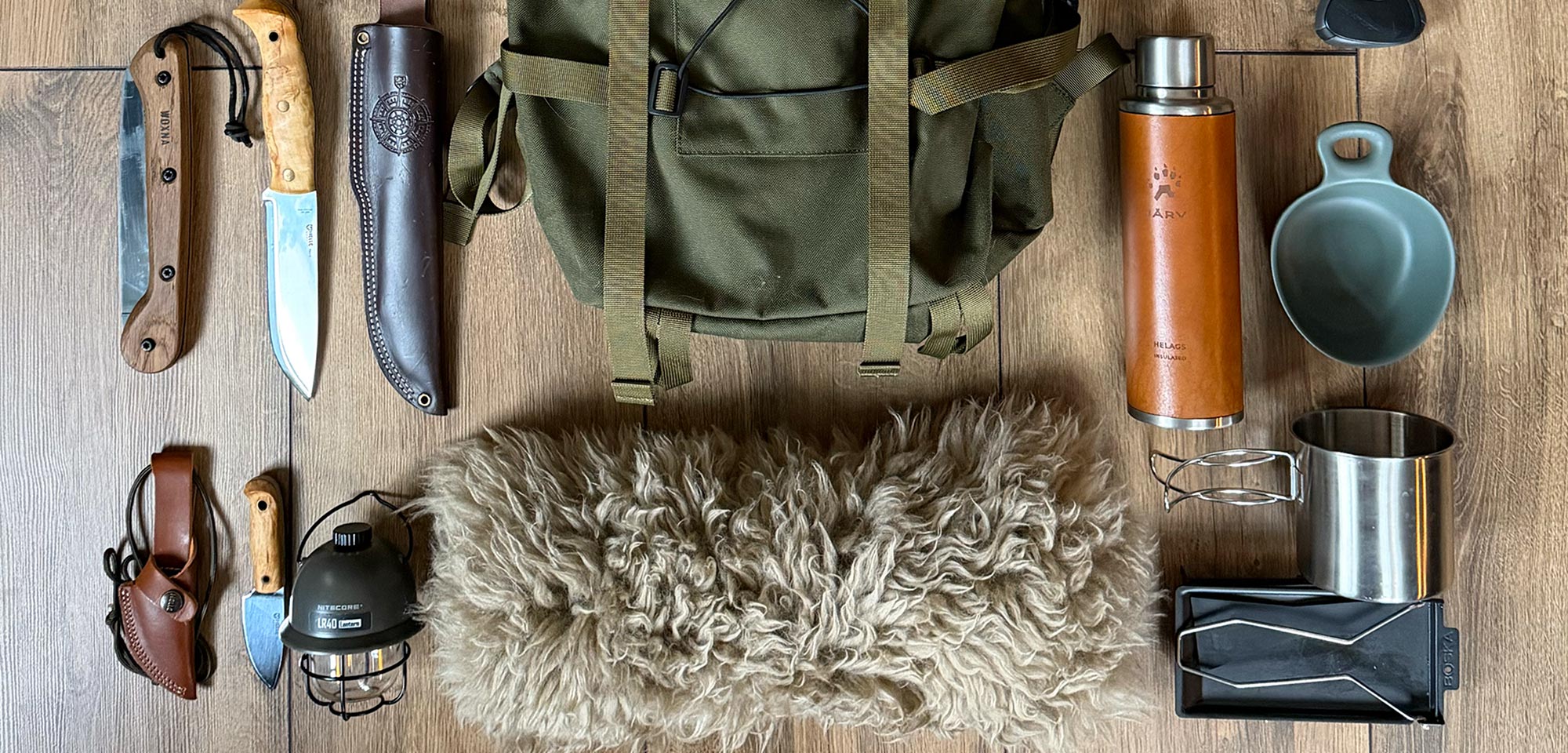 Flatlay mit Outdoor-Ausrüstung auf Holz, Rucksack, Axt, Bushcraftmesser, Feldflasche und Campinglaterne