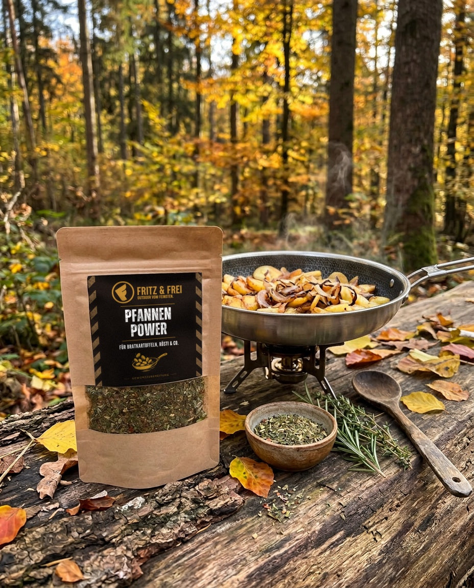 Fritz & Frei Pfannenpower Gewürzzubereitung 50 g im Kraftpapierbeutel auf einem Baumstamm im Herbstwald, daneben eine Pfanne mit Bratkartoffeln auf dem Campingkocher und eine Schale mit der Kräutermischung (KI)