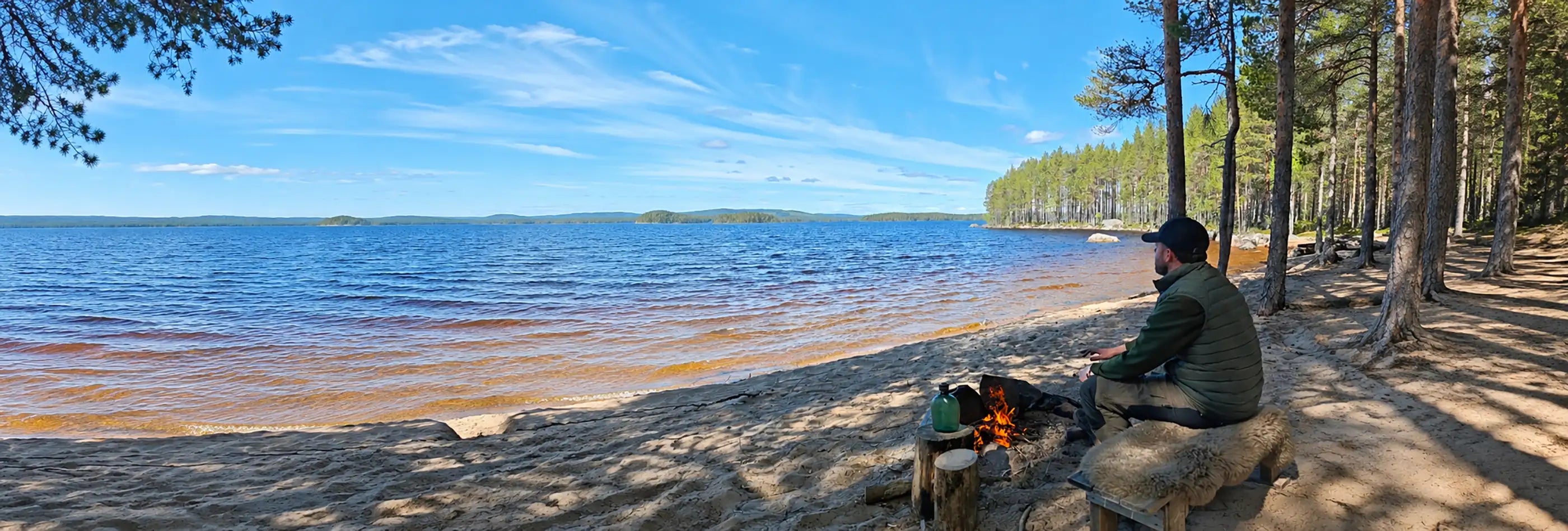 Mann sitzt mit Merino Schaffell am Lagerfeuer an einem See in Schweden