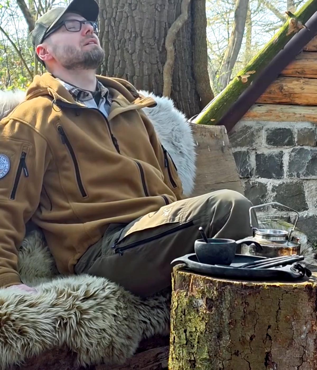 Merino Schaffell L in Cappuccino als Sitzunterlage am Baumstumpf im Wald, daneben Kessel und Kochgeschirr