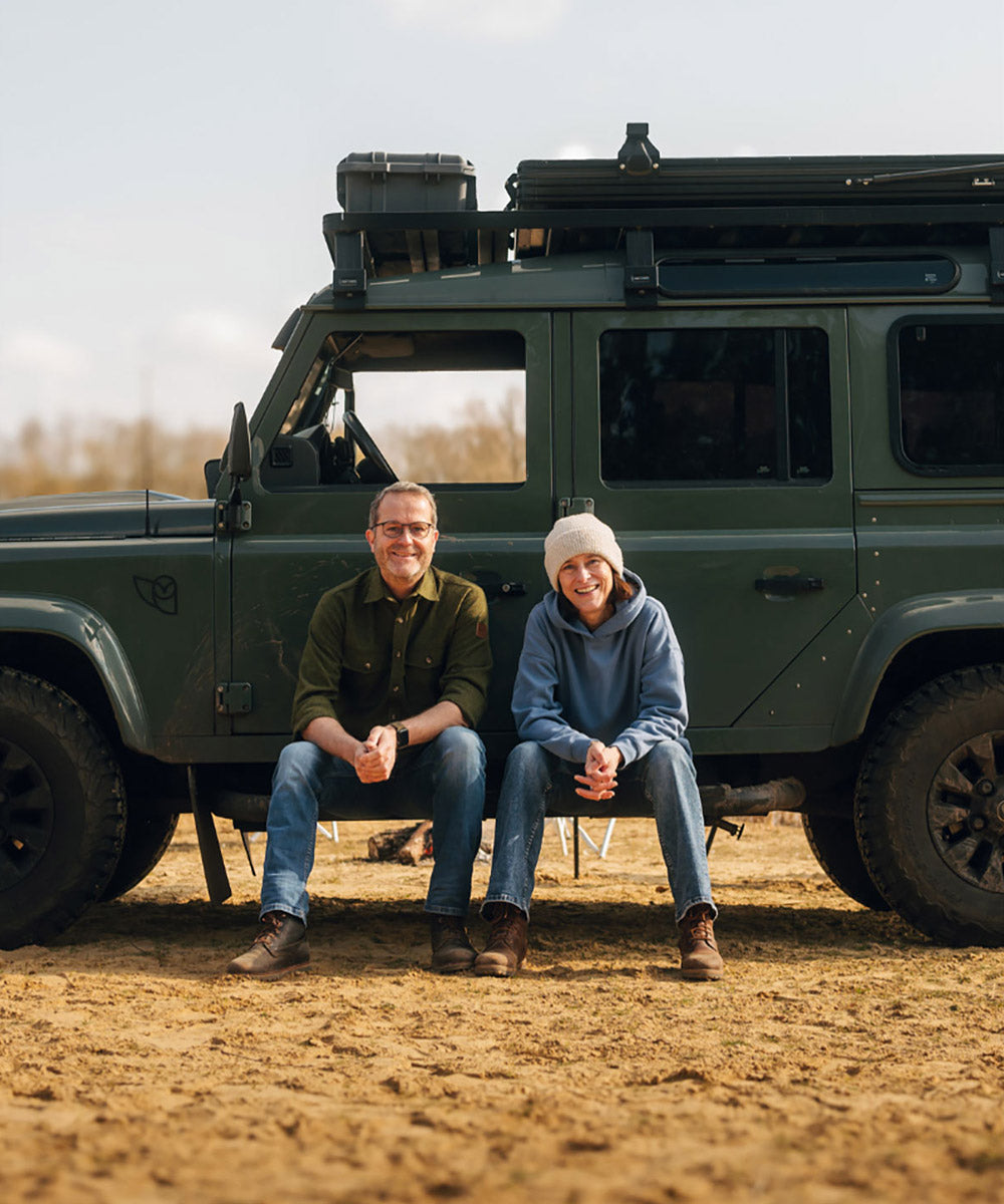 Lars und Kerstin von Fritz & Frei sitzen auf Campingstühlen vor ihrem grünen Geländewagen